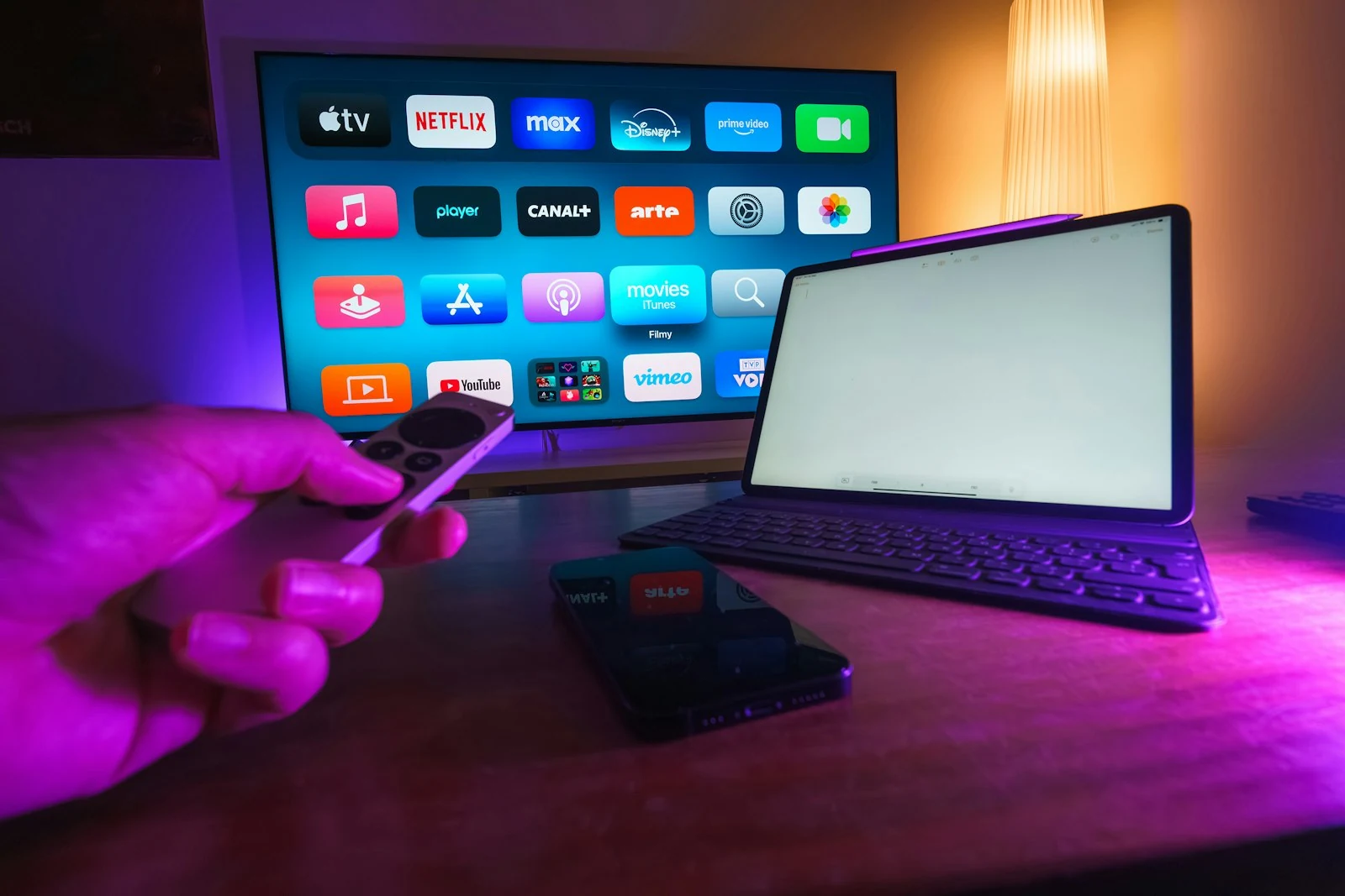 Une main tient une télécommande devant un écran affichant des applications de streaming, à côté d’une tablette.